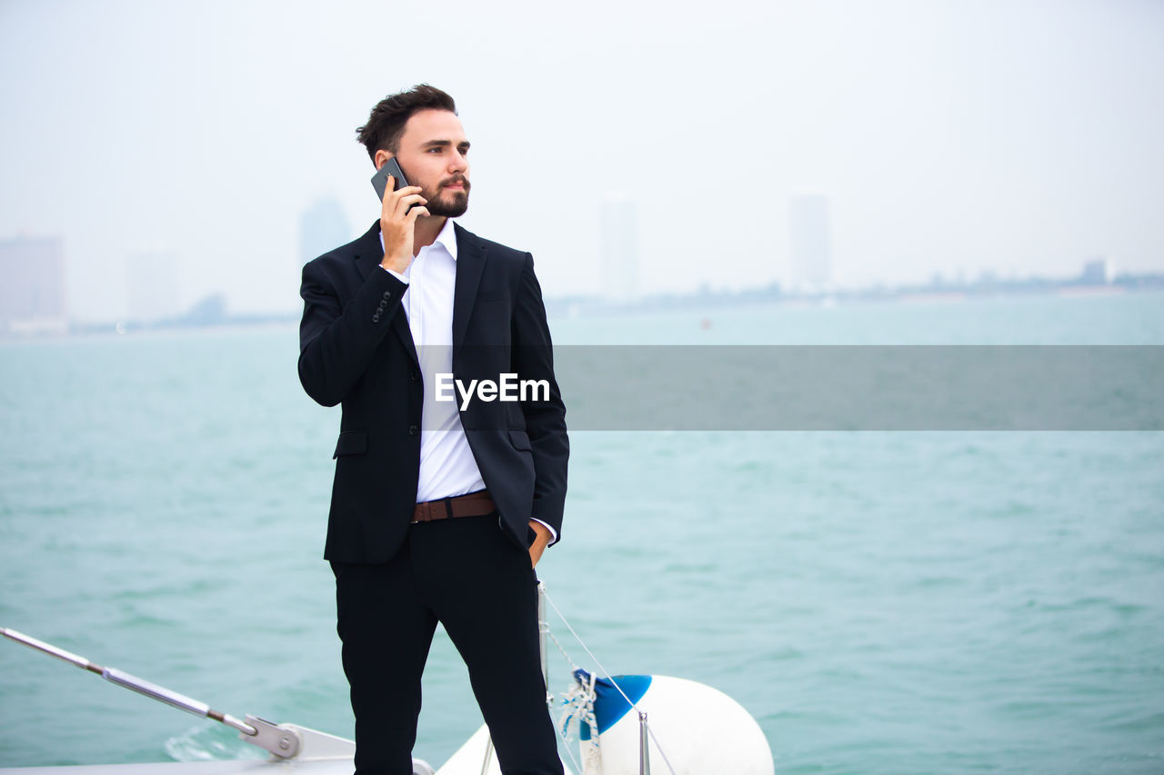 Businessman talking on phone while standing on boat in sea against clear sky