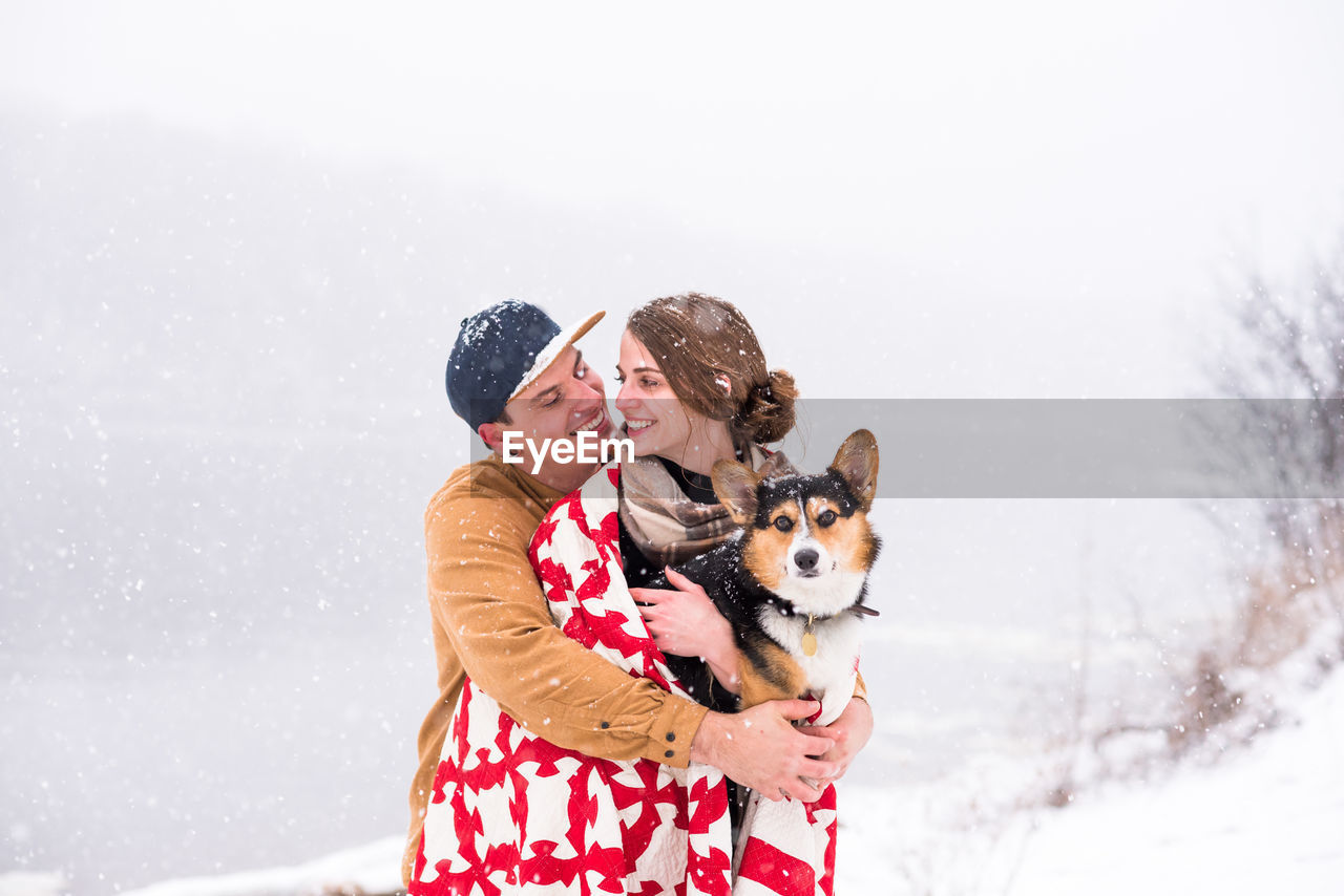 Young couple smile and huddle during snowfall with dog