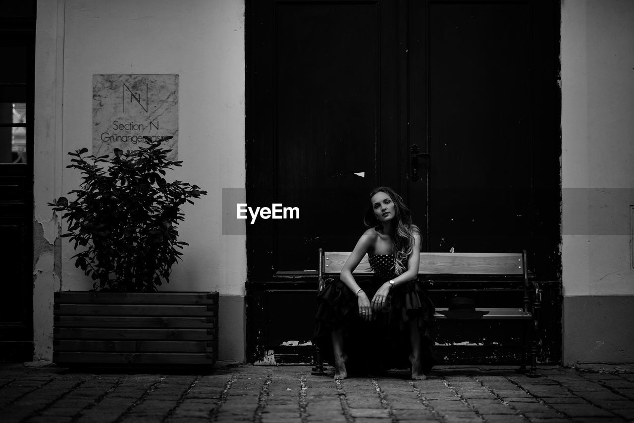 FULL LENGTH PORTRAIT OF WOMAN SITTING ON SEAT AGAINST WALL