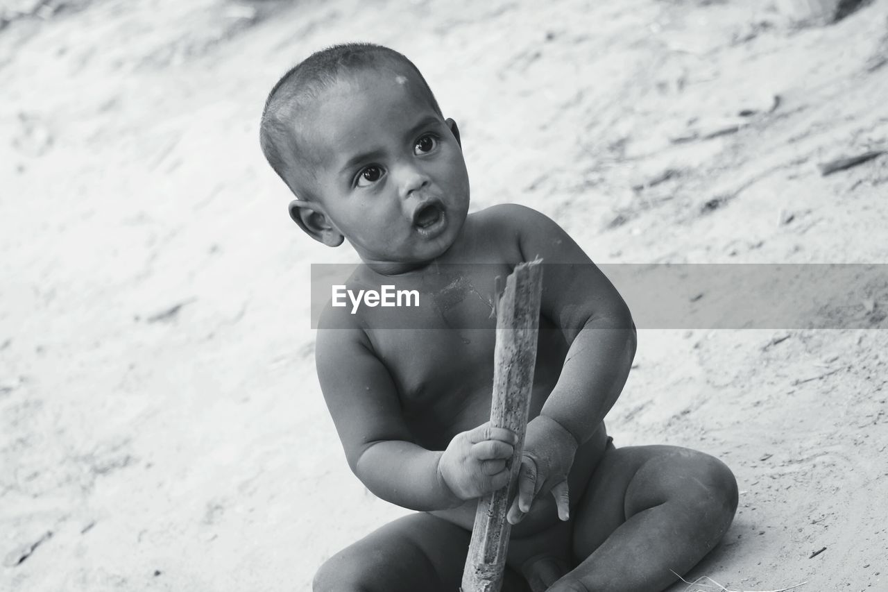 Naked baby boy holding wood while sitting on sand at beach