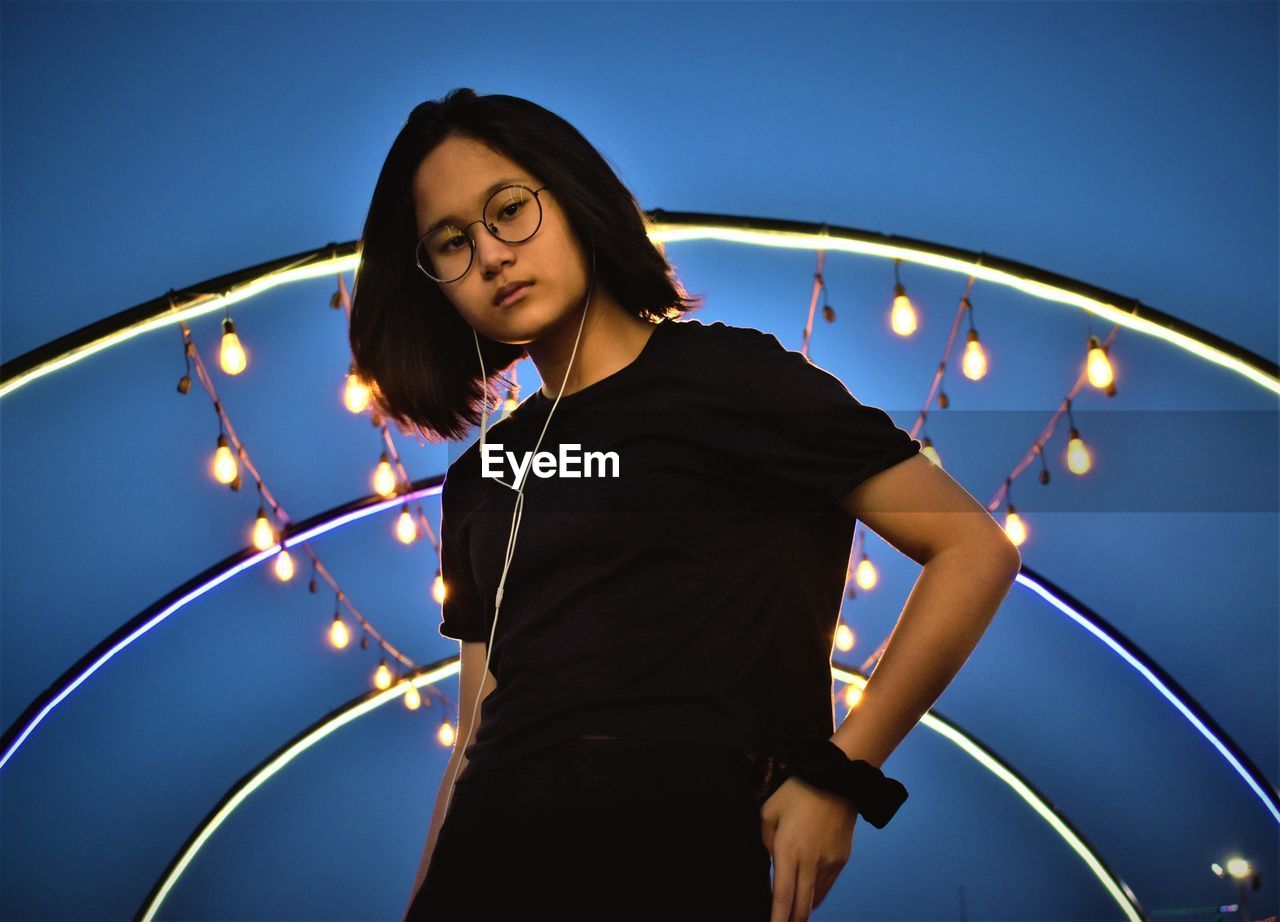 Young woman looking away while standing against illuminated lights