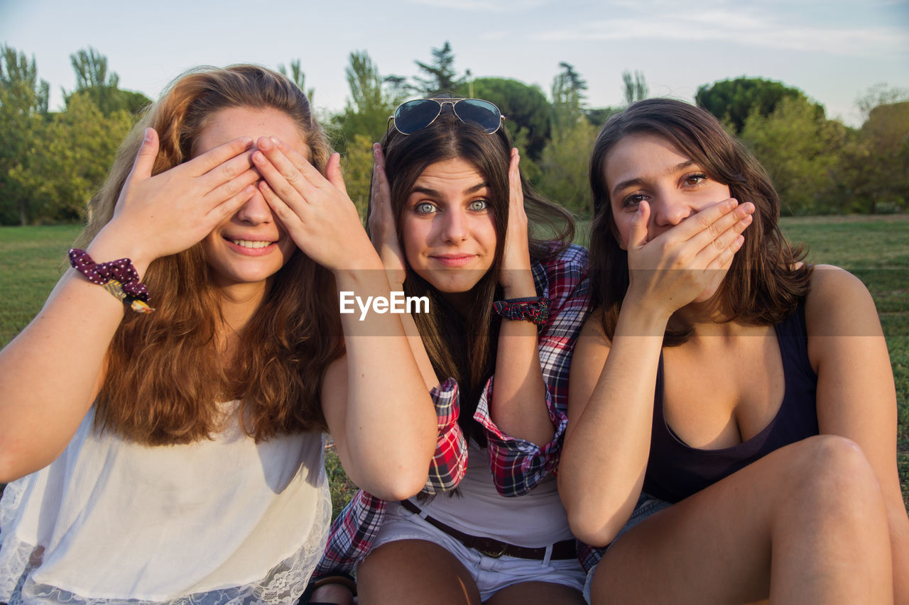 Young women imitating see no evil hear no evil speak no evil while sitting on field