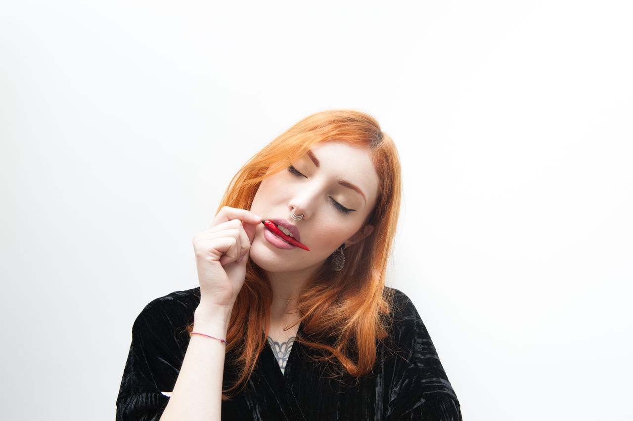 Close-up of woman with eyes closed holding red chili pepper in mouth against white background