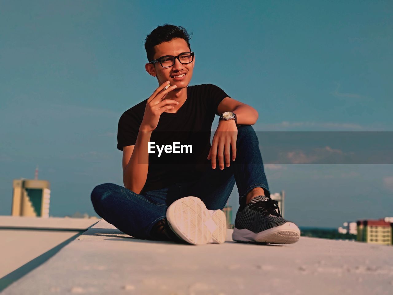 Smiling young man smoking cigarette while sitting on building terrace against blue sky