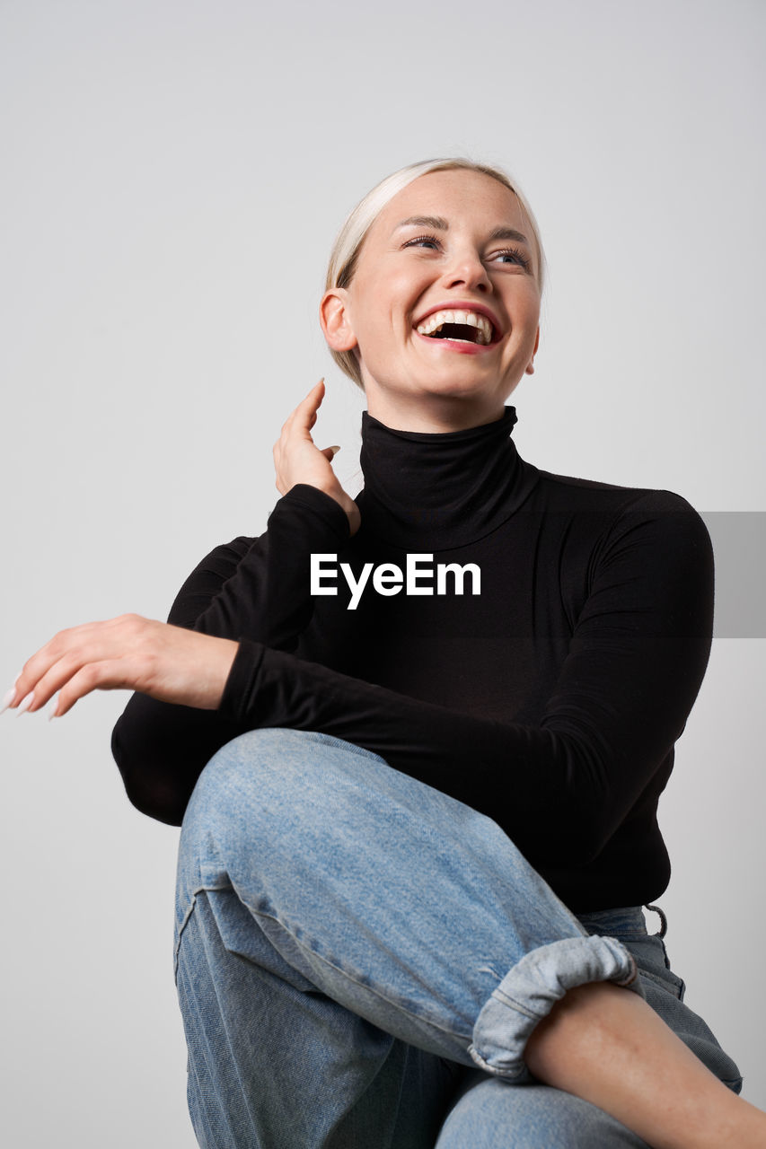 Cheerful blonde female in casual apparel sitting in studio and laughing sincerely while looking away