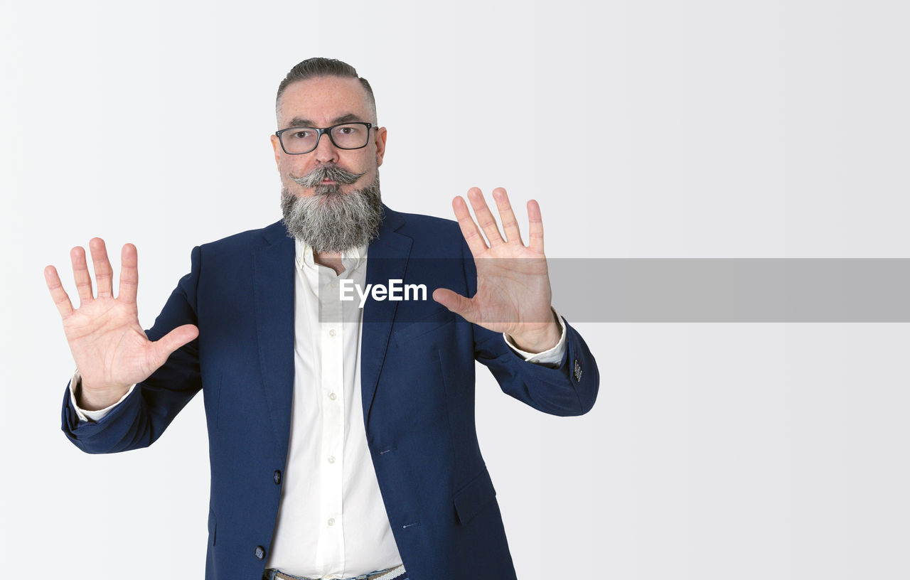 Stylishly dressed bearded hipster in blue jacket on white, raising hands and showing palms,
