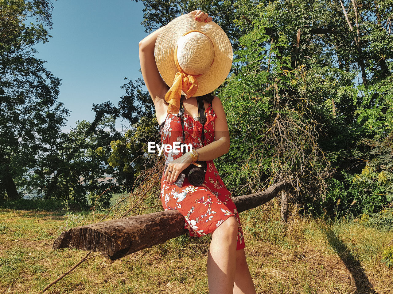 Woman covering face with hat while sitting on tree trunk