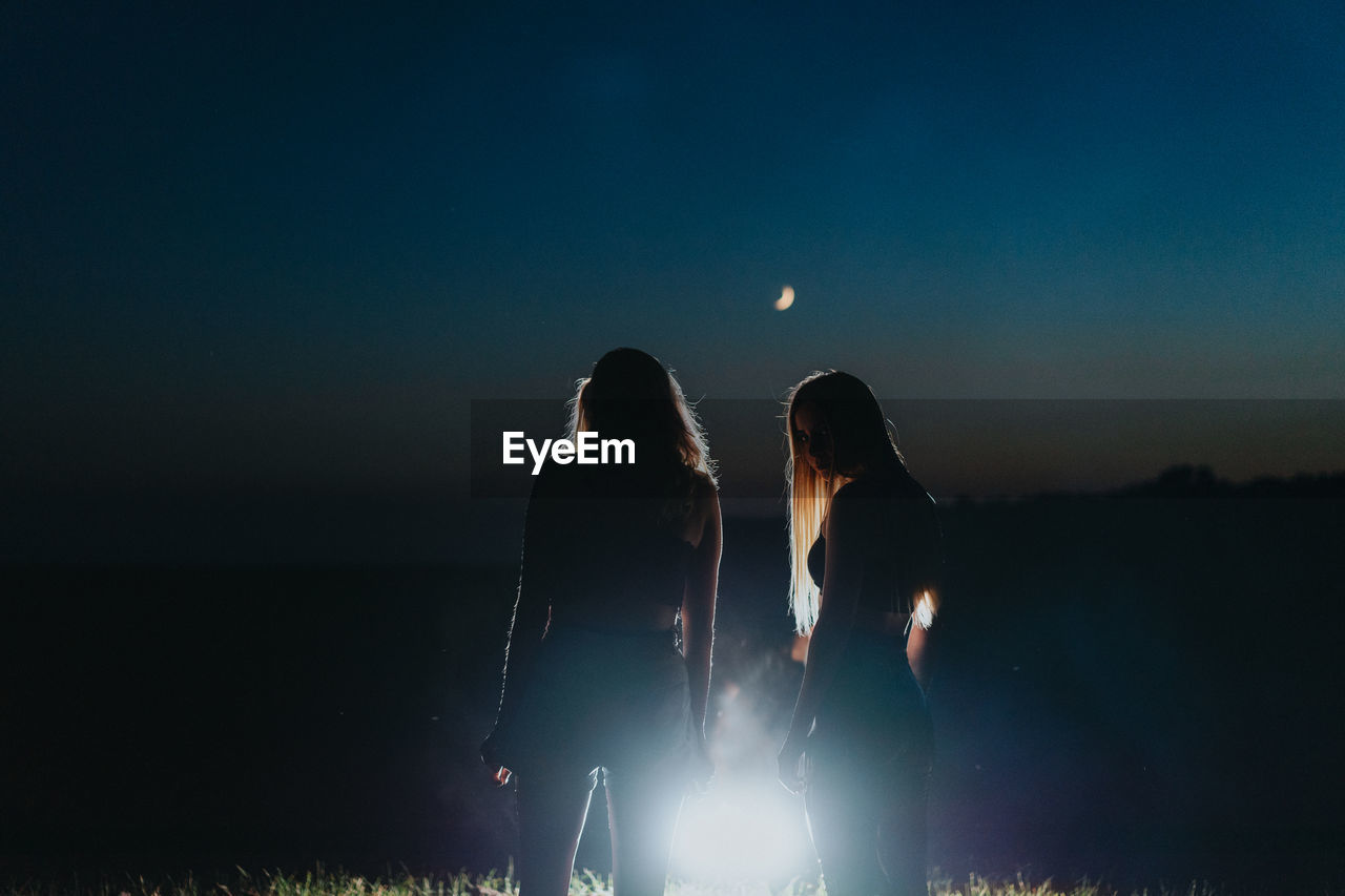 Silhouette women standing against illuminated background against sky during dusk