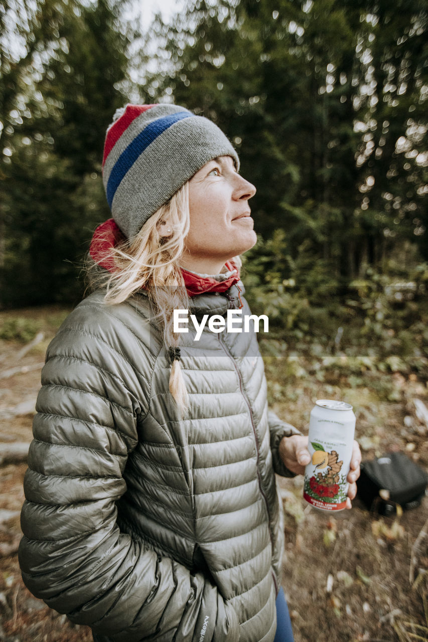 Smiling happy blonde woman in gray jacket and winter hat holding beer.