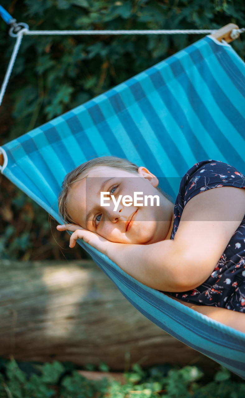 A girl with blond hair and in a dress lies in a hammock outdoors on a sunny summer day