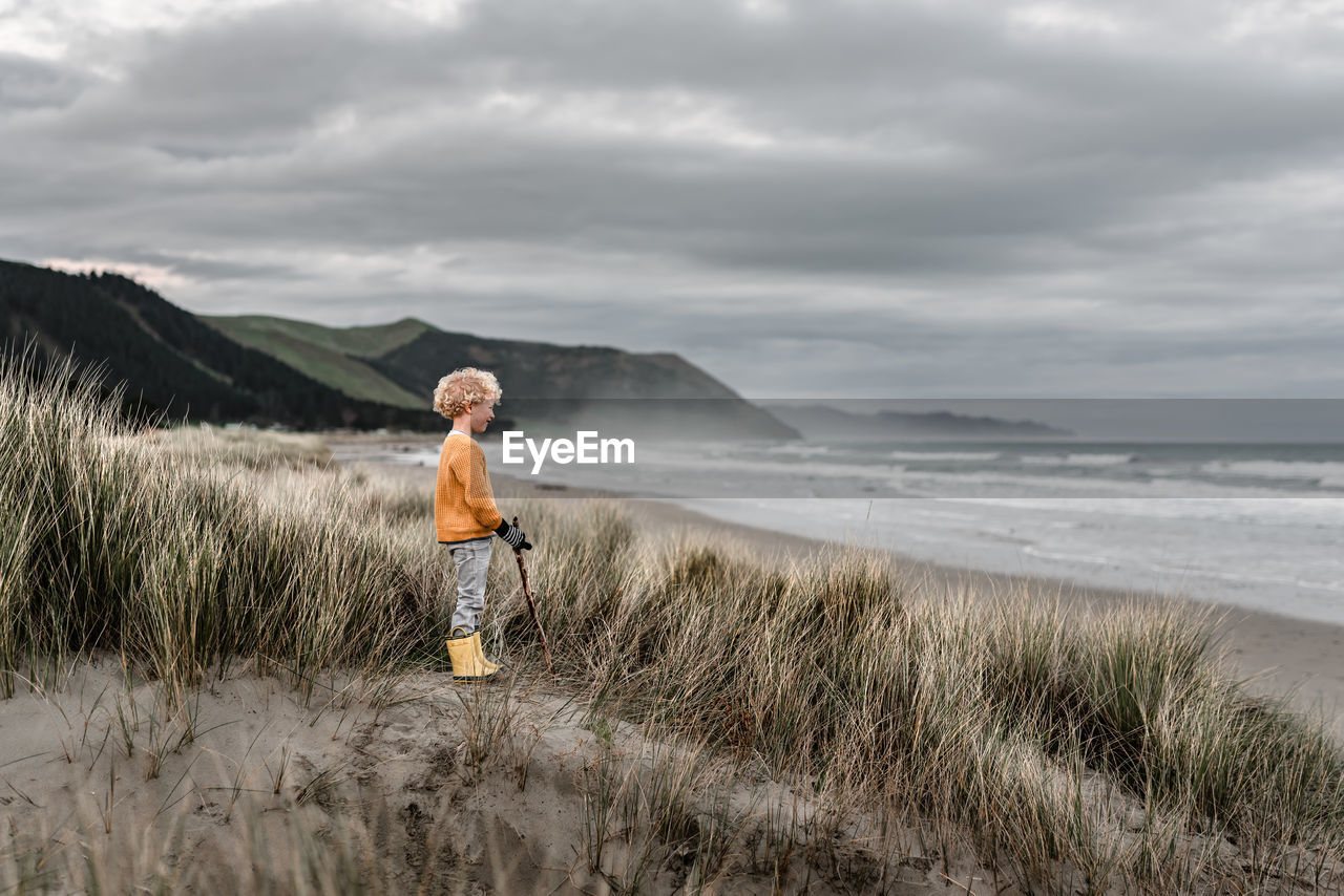 Blond curly haired boy watching ocean on cloudy day in new zealand