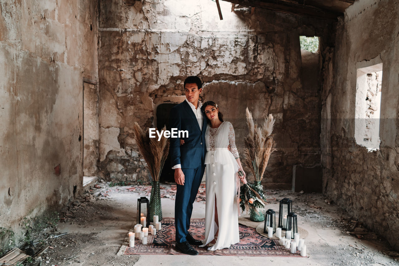 Side view full body loving young ethnic newlywed couple wearing chic wedding clothes embracing and kissing gently in obsolete remaining building