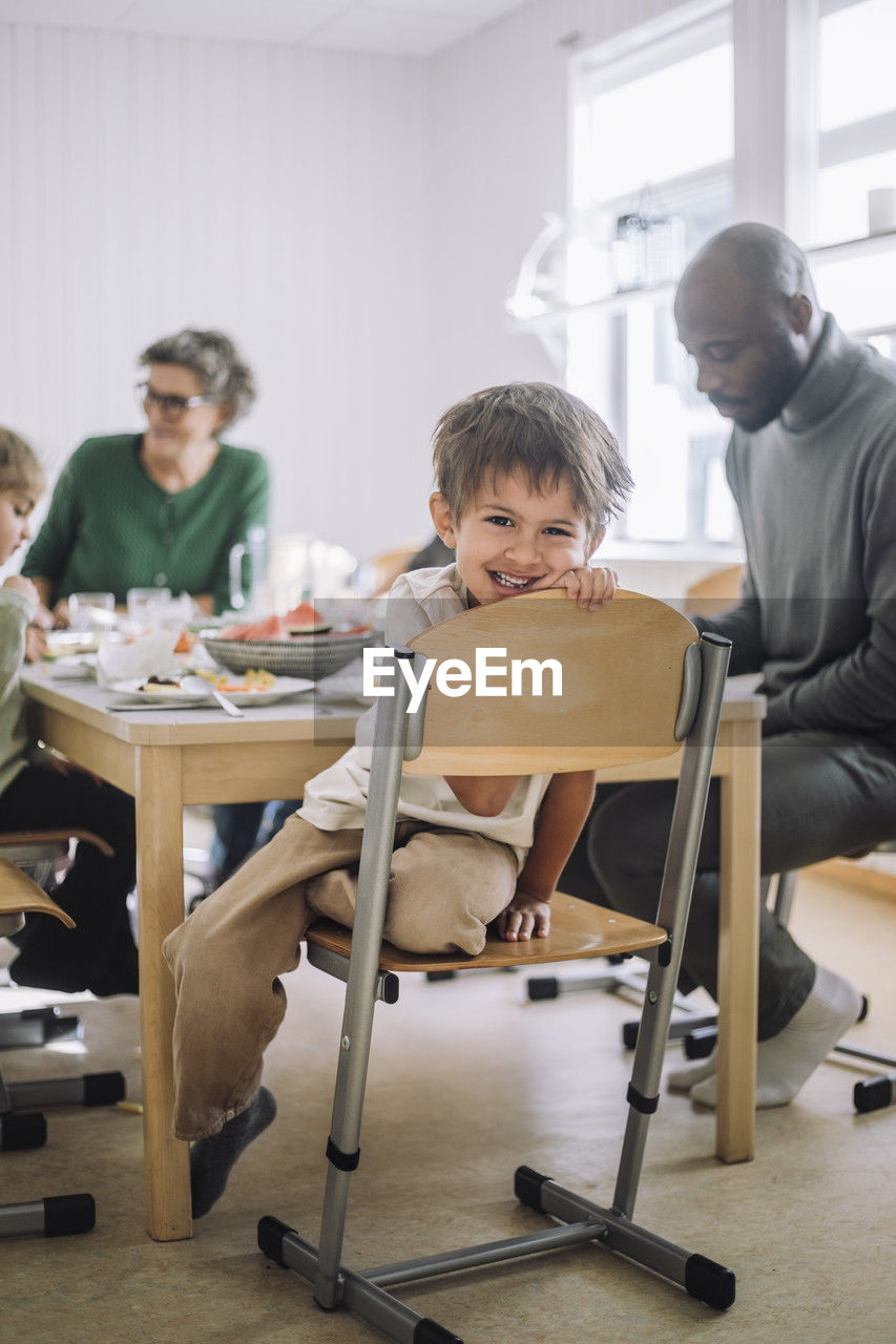 Portrait of smiling boy sitting on chair with teacher at dining table during lunch break
