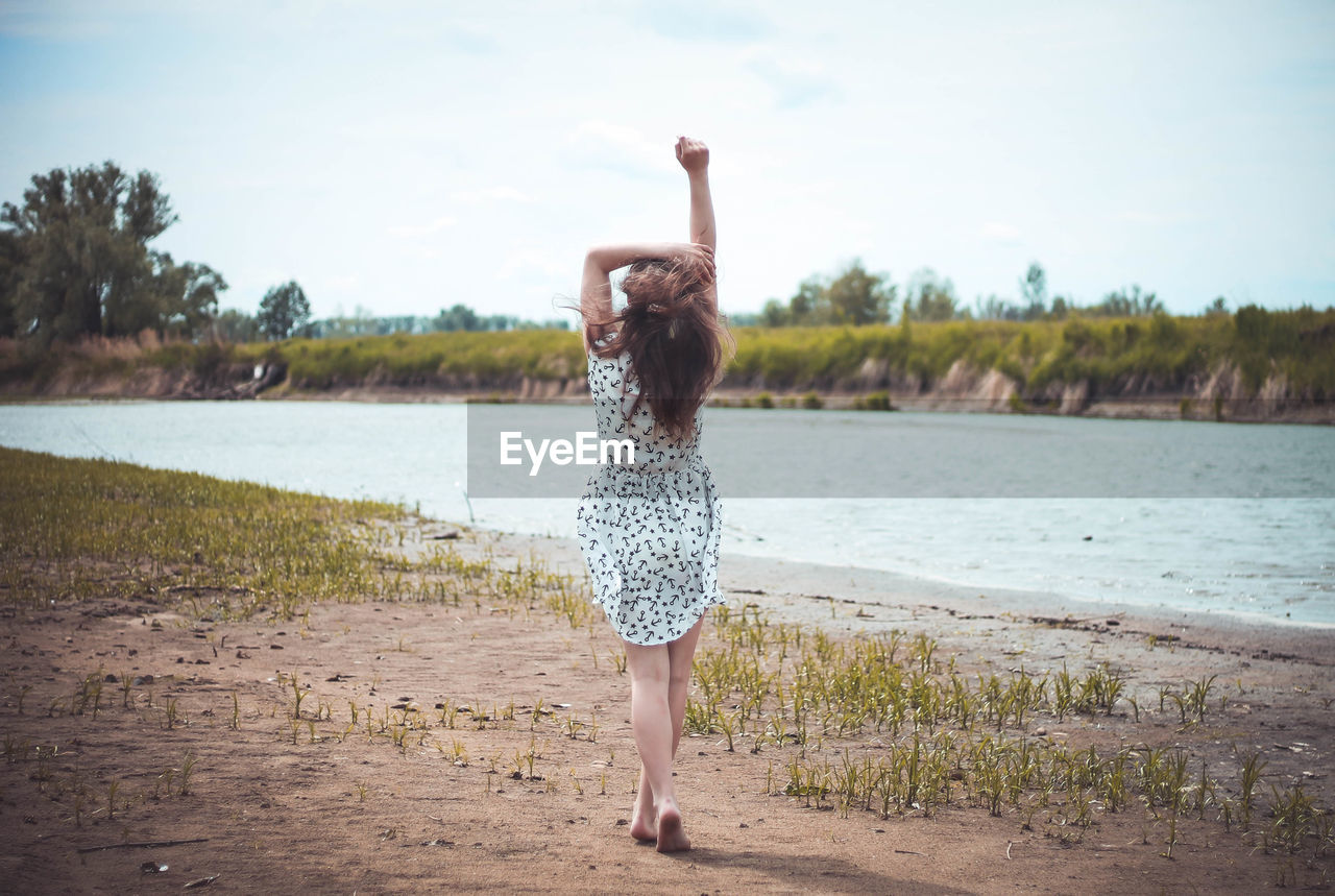 Full length of woman walking with arms raised by lake against sky