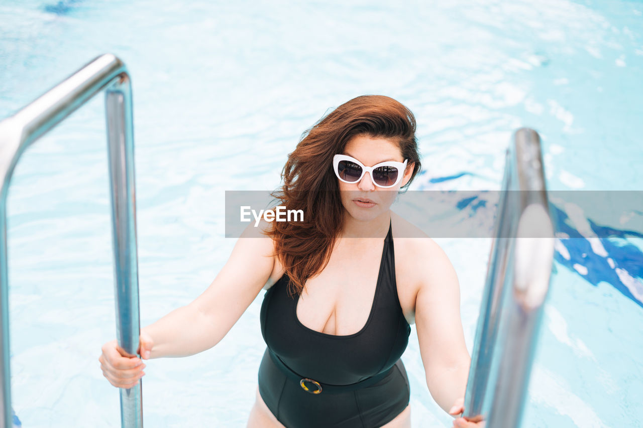 Stylish woman plus size body positive in black swimsuit and white sunglasses swimming in hotel pool