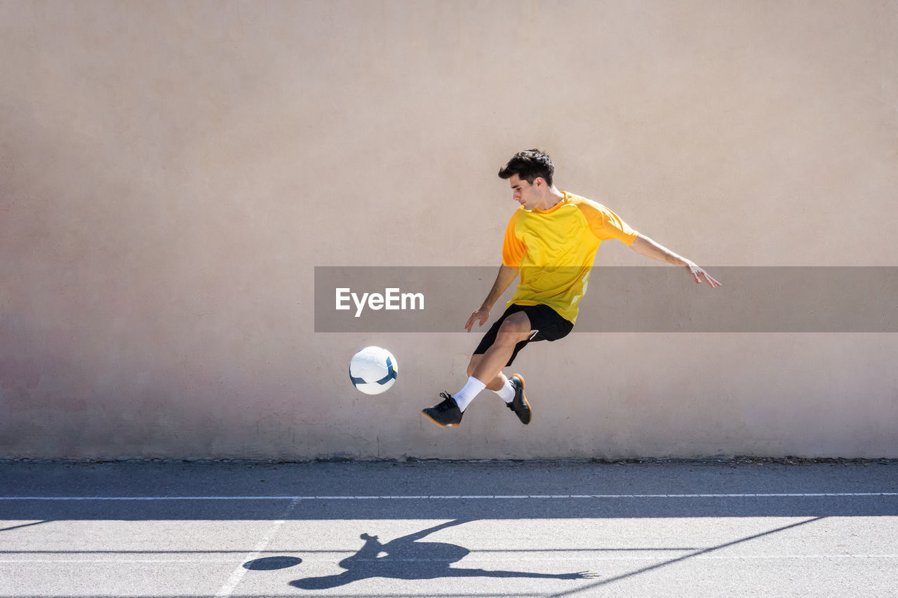 Young male player kicking football while practicing soccer against building