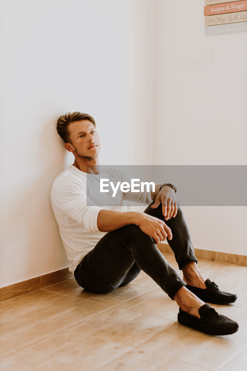 Full length of man looking away while sitting on hardwood floor against wall