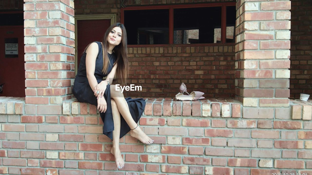 PORTRAIT OF WOMAN SITTING AGAINST BRICK WALL