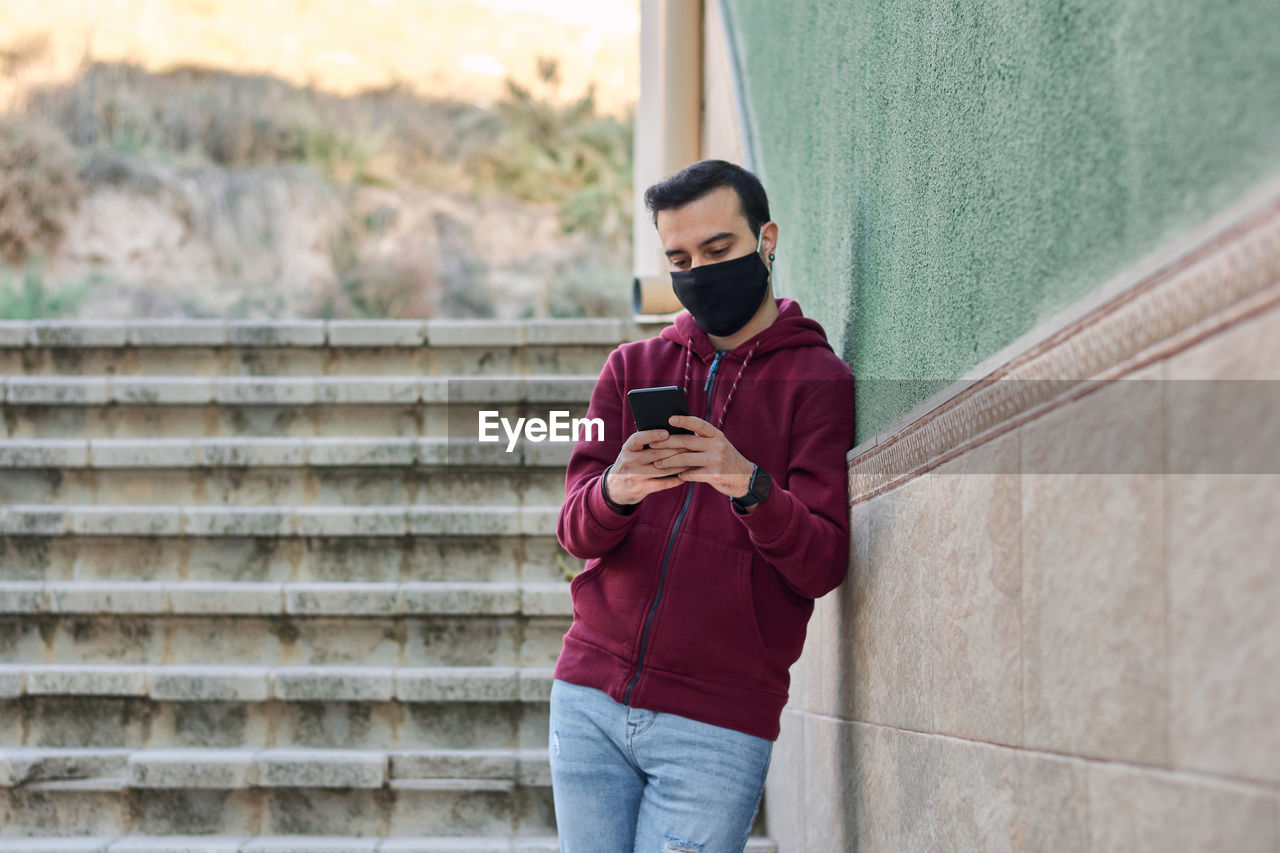 Young man with a mask writes on his phone leaning against a wall