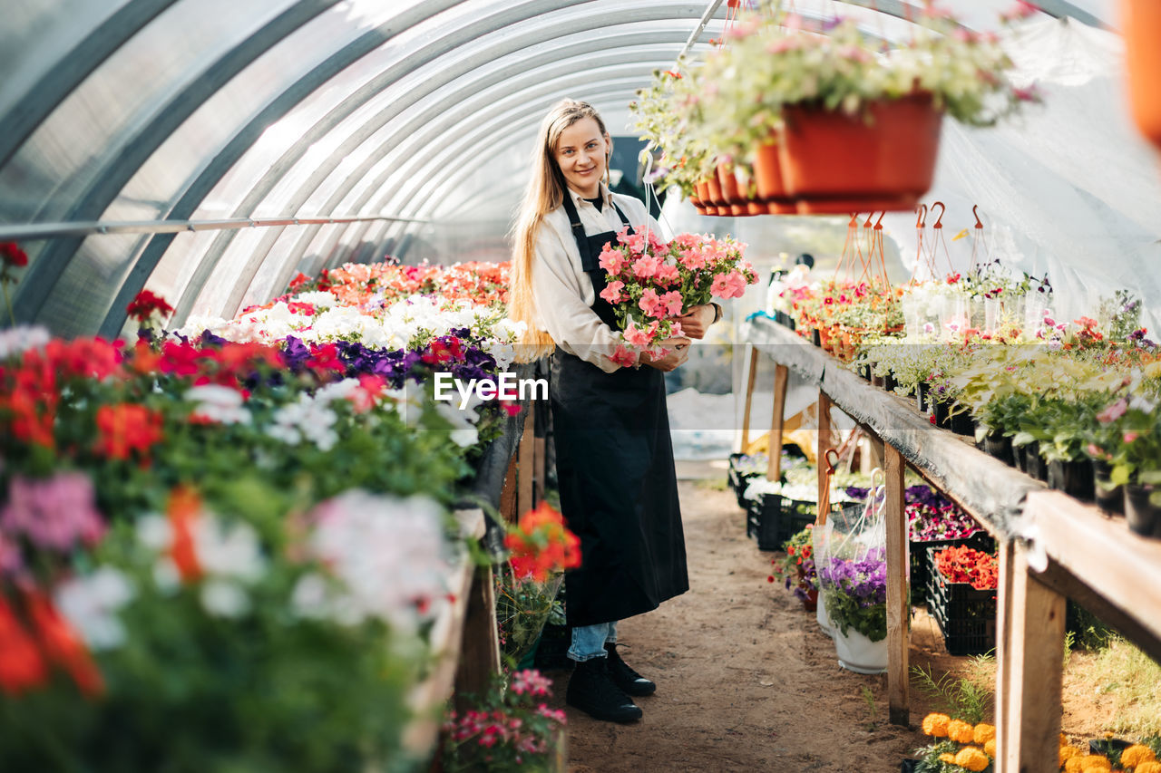 A cheerful woman in an apron holds a pot of flowers in her hands while working in a greenhouse