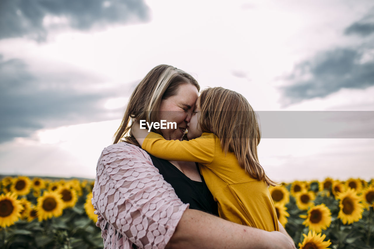 Mother and daughter embracing while standing in sunflower farm against sky