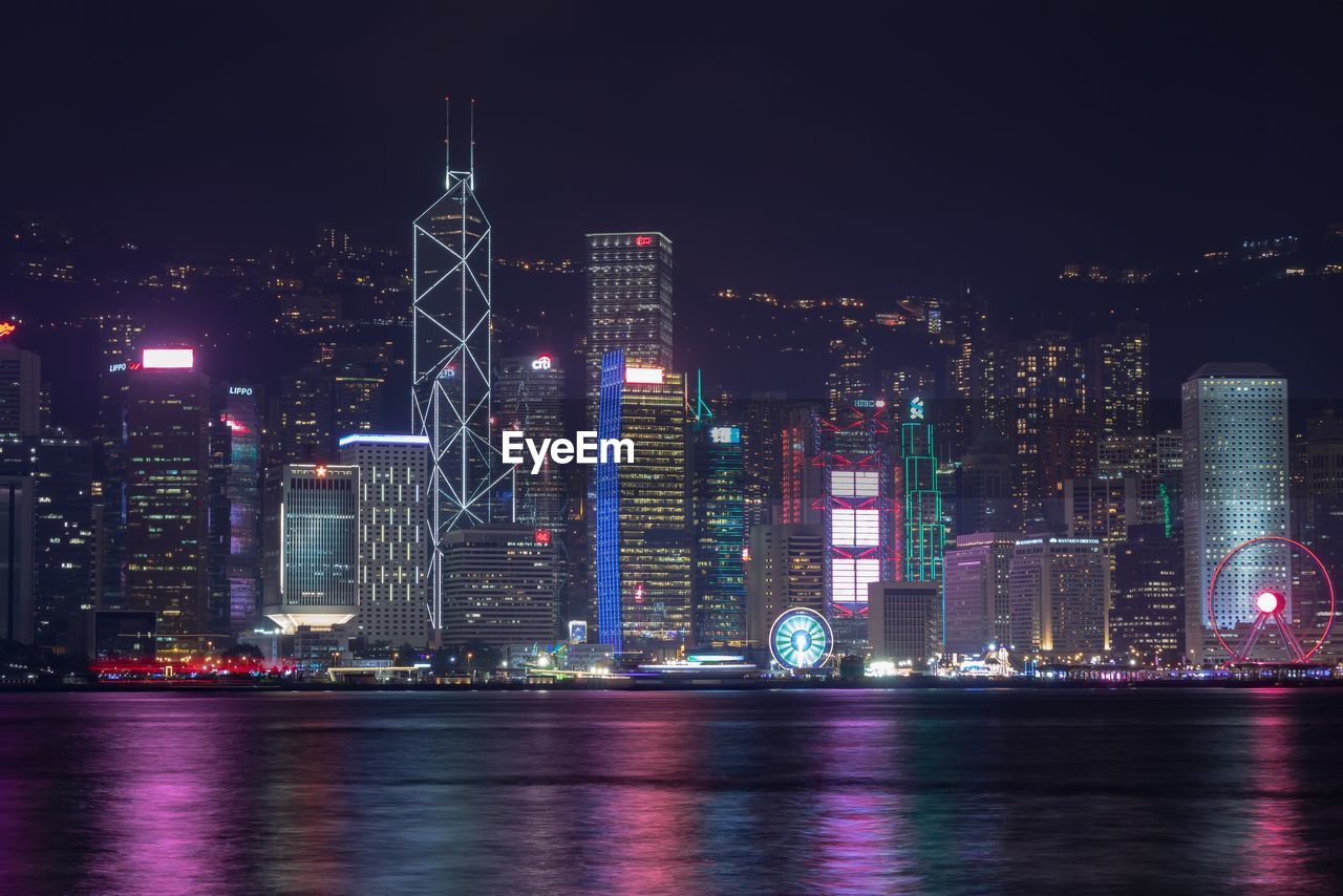 Illuminated modern buildings by river against sky at night