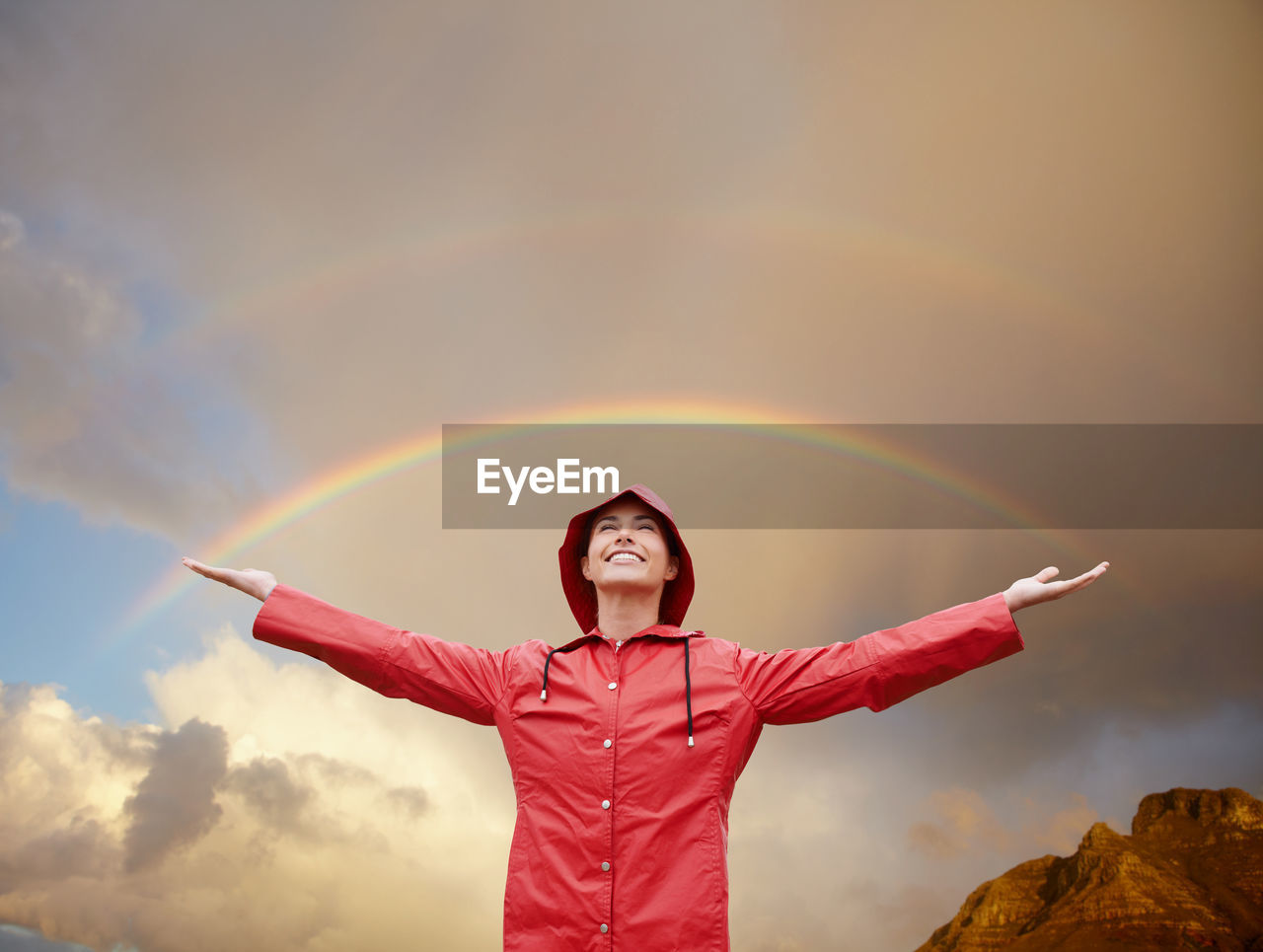 portrait of smiling young woman with arms raised standing against sky