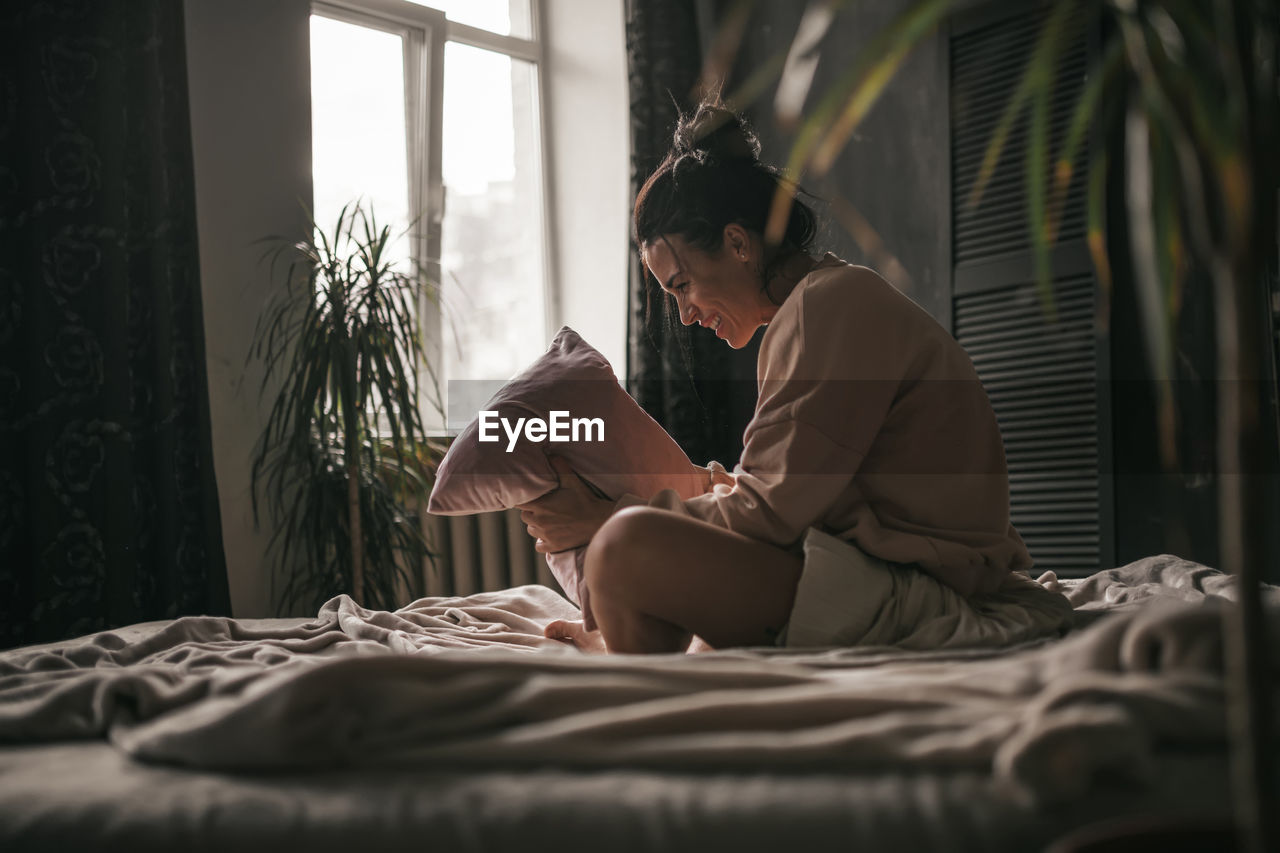 Side view of woman holding pillow while sitting on bed