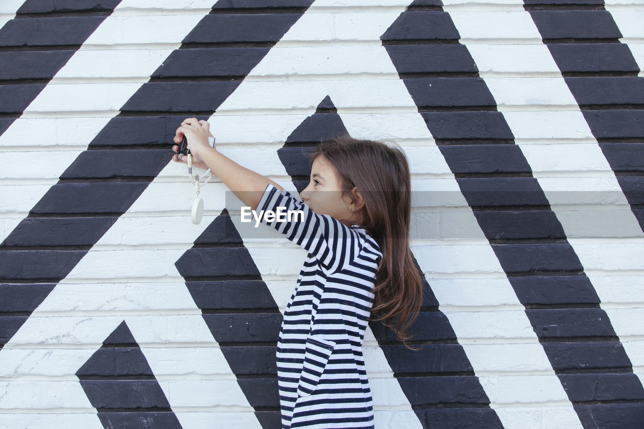 Side view of girl photographing with camera while standing against wall