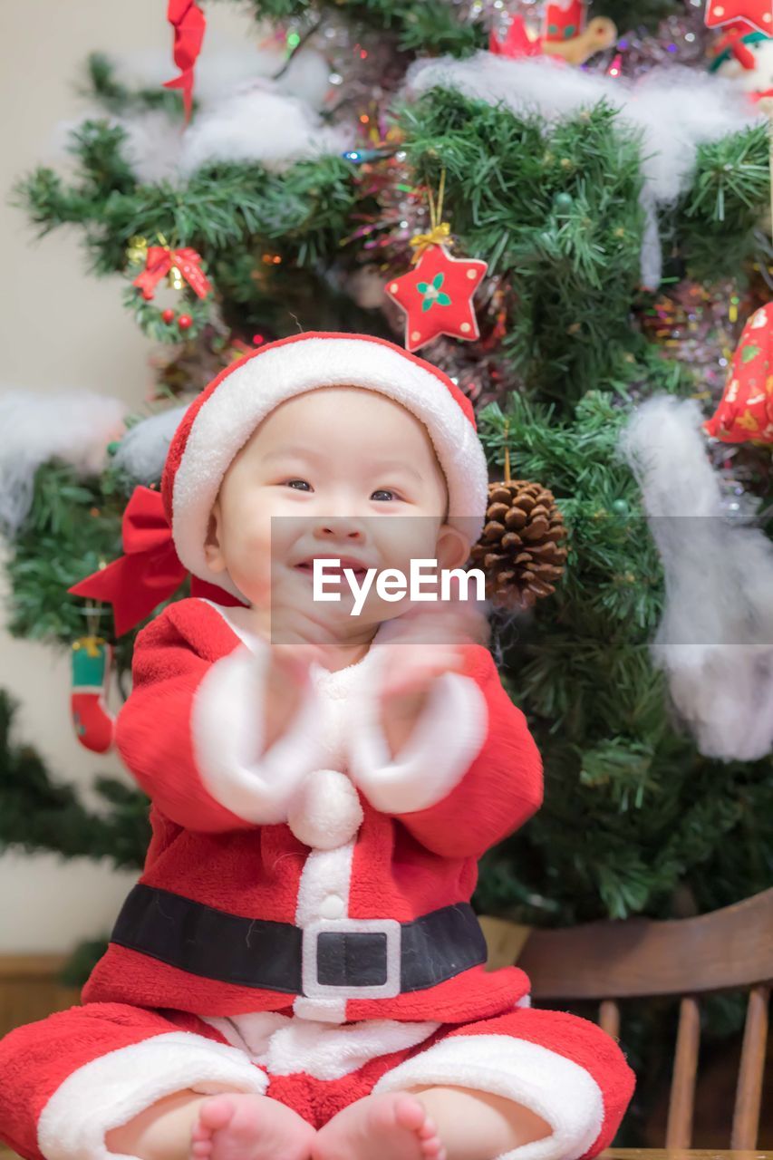 Cute baby girl wearing santa costume sitting against christmas tree