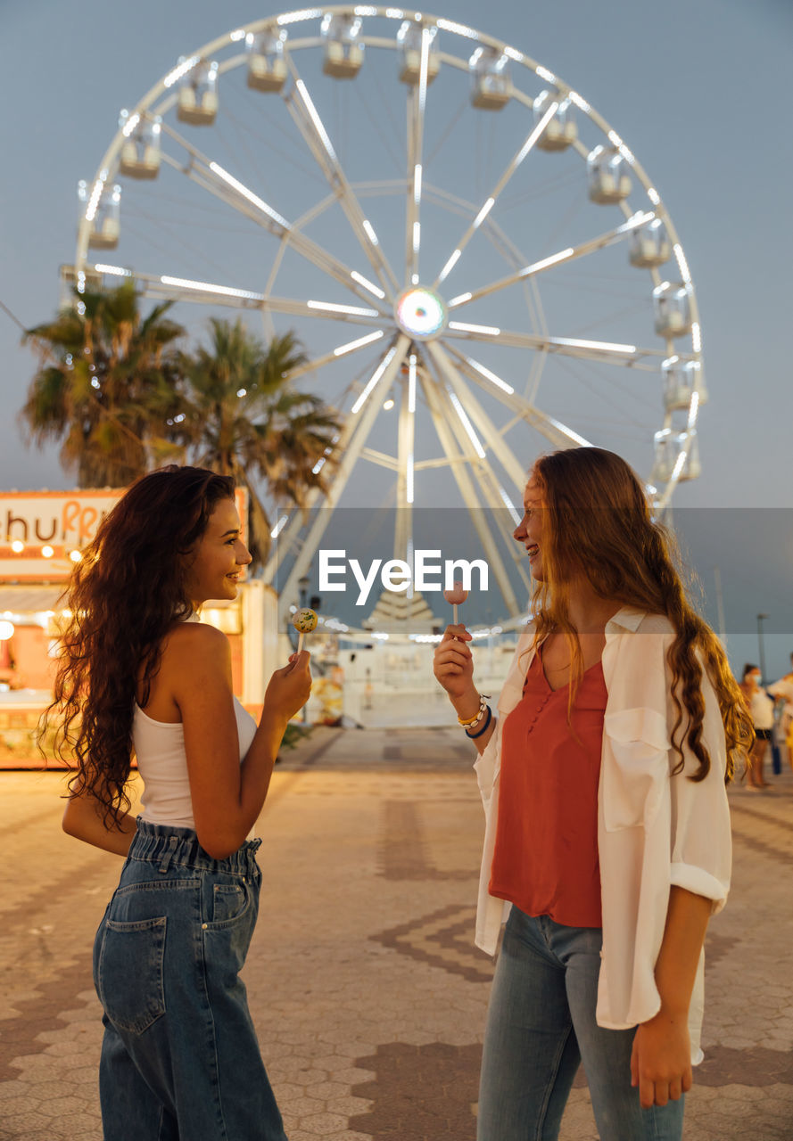 Low angle side view of happy carefree teen female friends with candies in hands enjoying summer evening on funfair while standing against ferris wheel