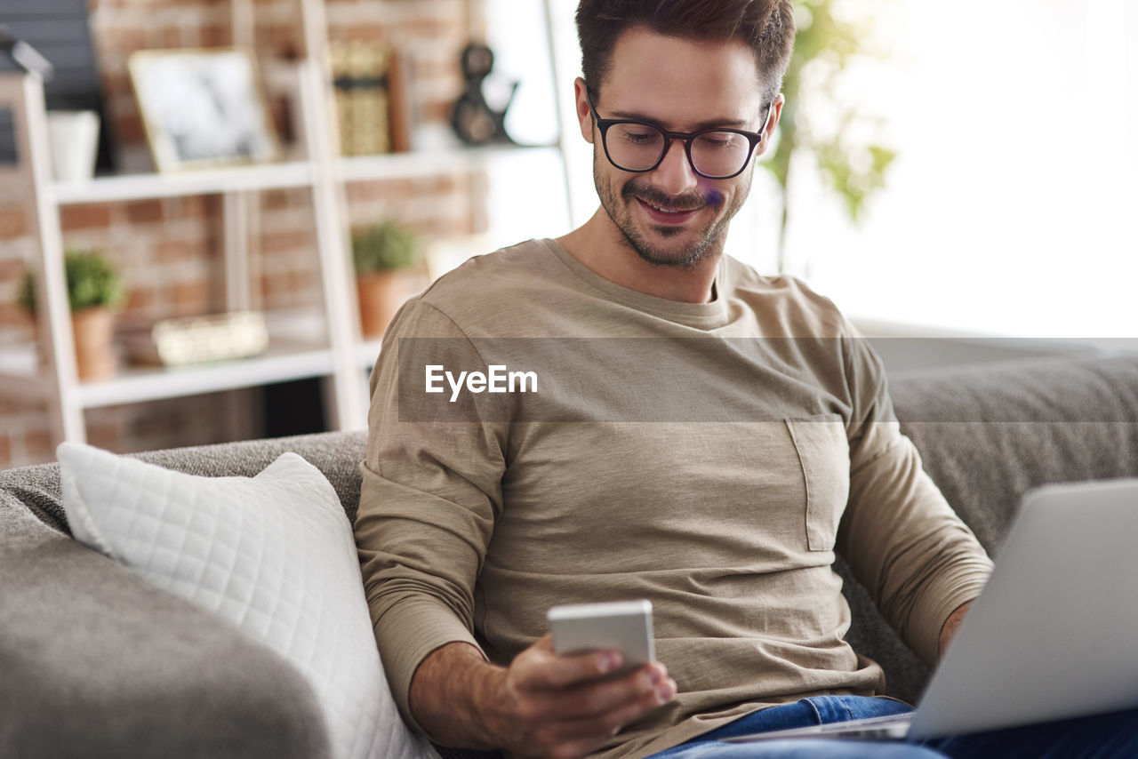 Smiling man sitting on couch at home using laptop and cell phone