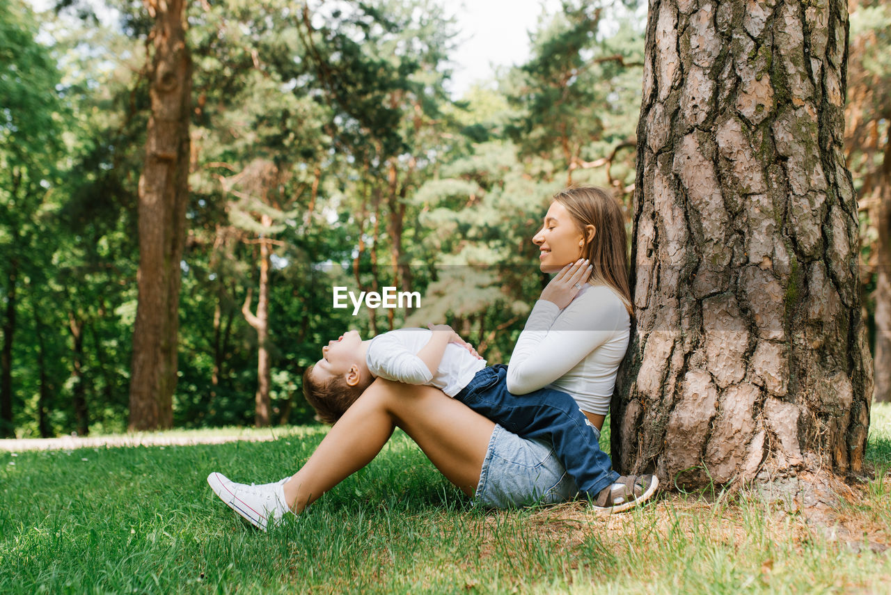 Young caucasian mom and her three-year-old son have fun sitting in the park near a tree on the grass