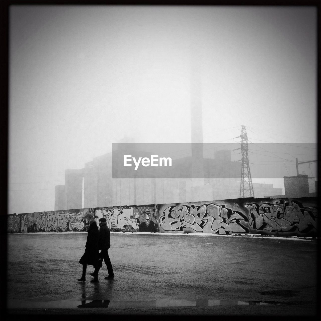 Couple walking on street by graffiti against factory covered with fog at dusk