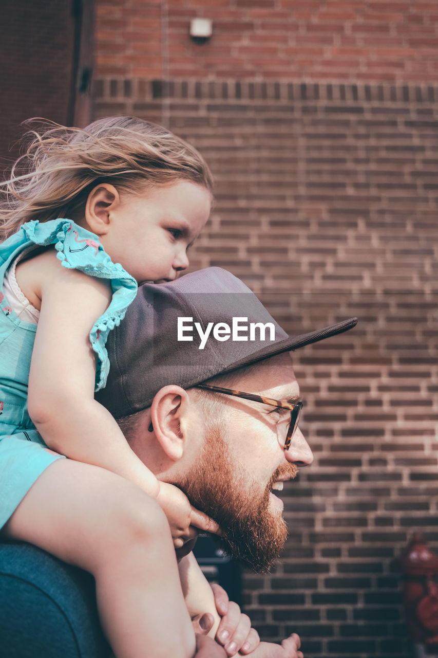 Side view of mid adult man carrying daughter on shoulders against brick wall