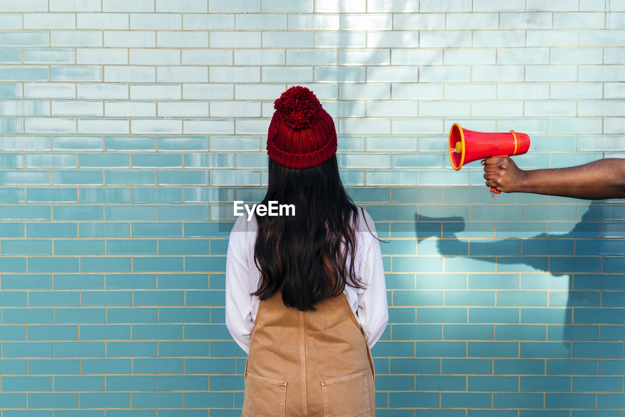 Man's hand holding megaphone by woman facing turquoise brick wall