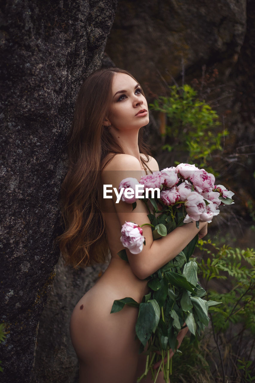 Naked woman holding bouquet while standing against tree