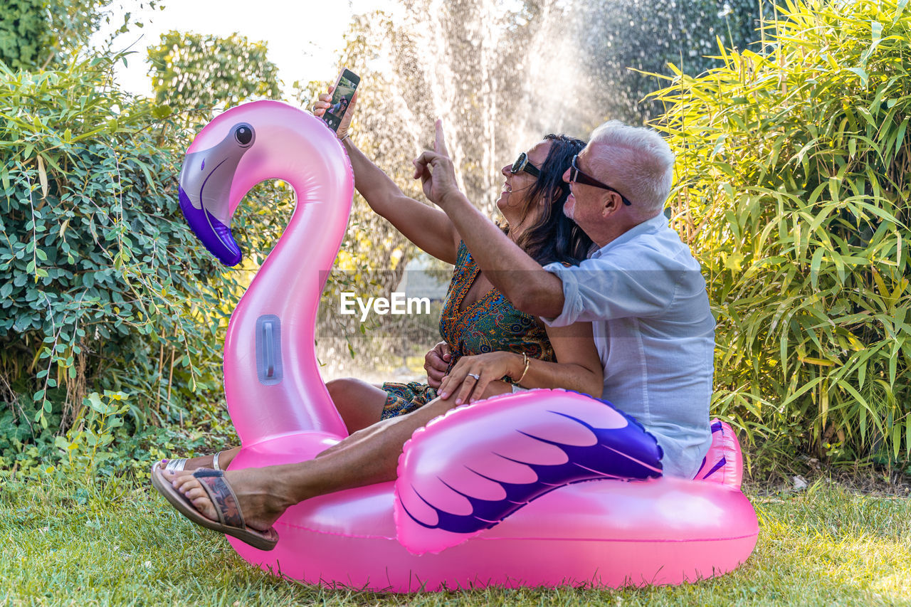 Beautiful smiling middle aged couple having fun taking a selfie sitting on flamingo inflatable toy