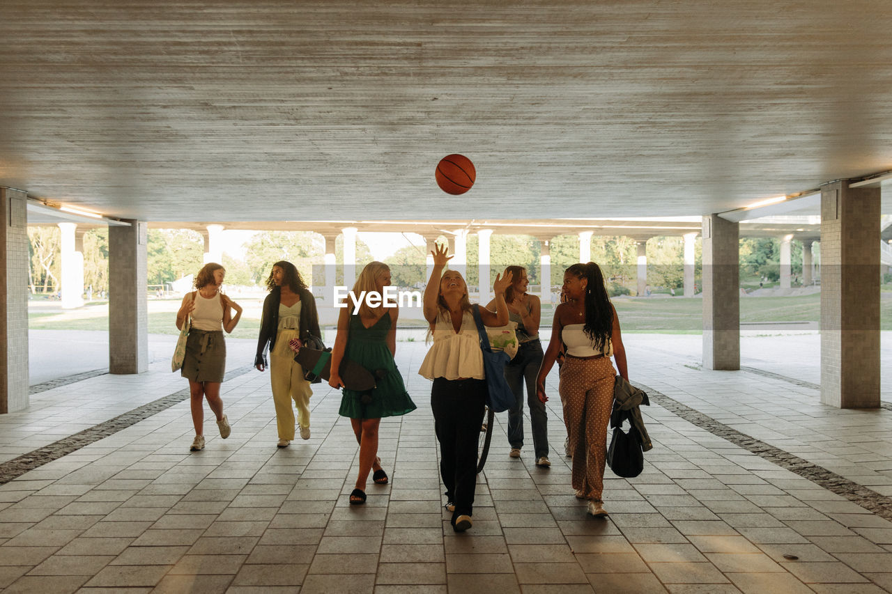 Teenage girl throwing basketball while walking with female friends under bridge