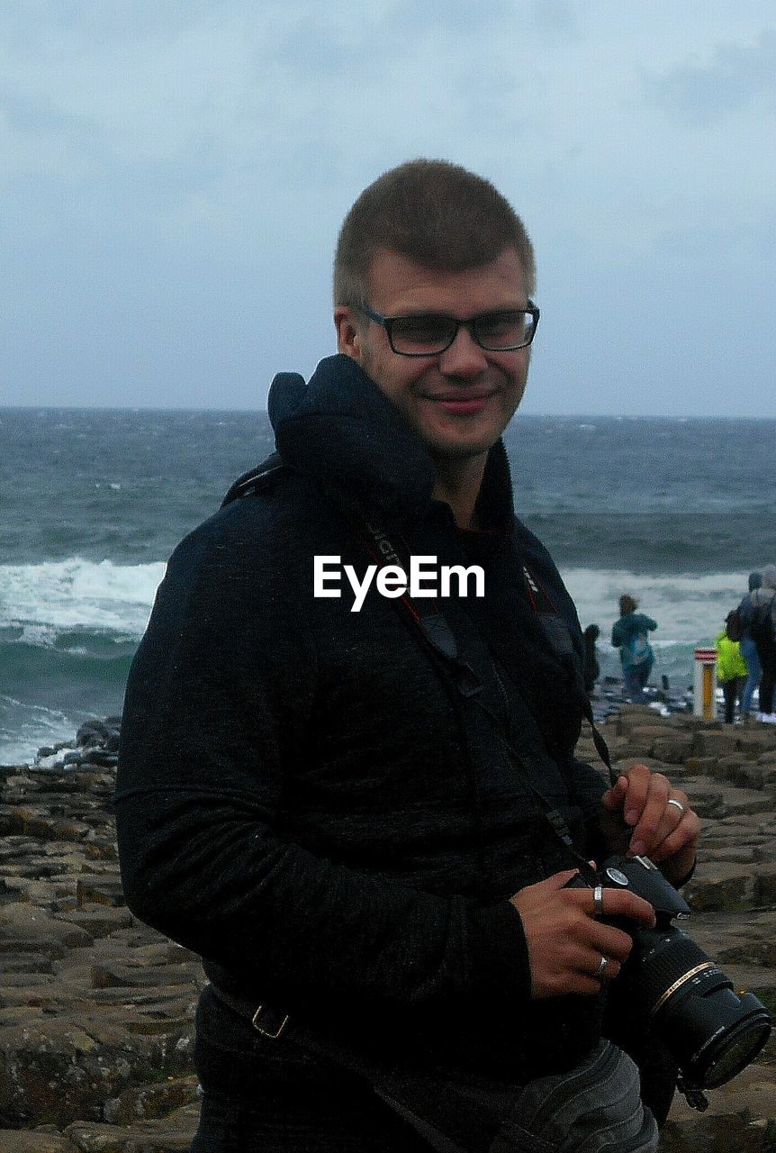 PORTRAIT OF SMILING MAN STANDING AT BEACH