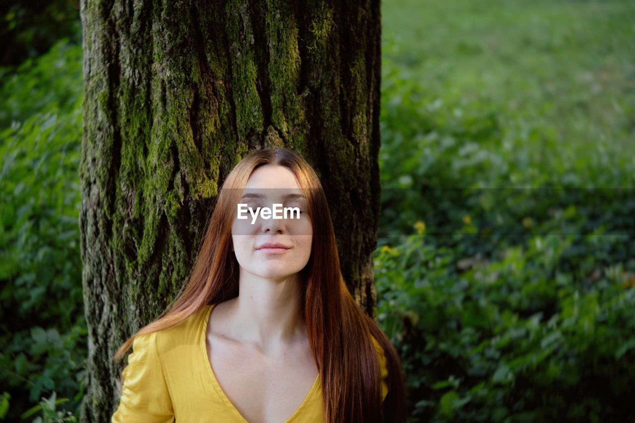 Young woman with eyes closed standing by tree trunk