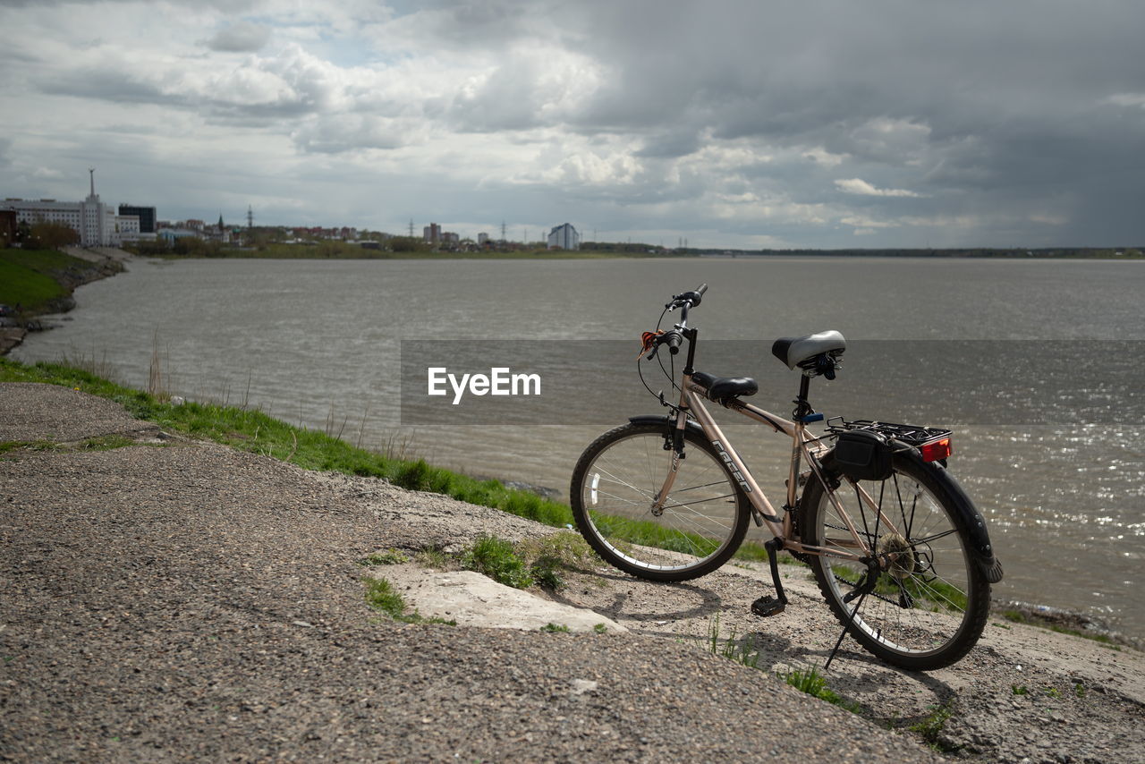 BICYCLE PARKED ON ROAD BY RIVER AGAINST SKY