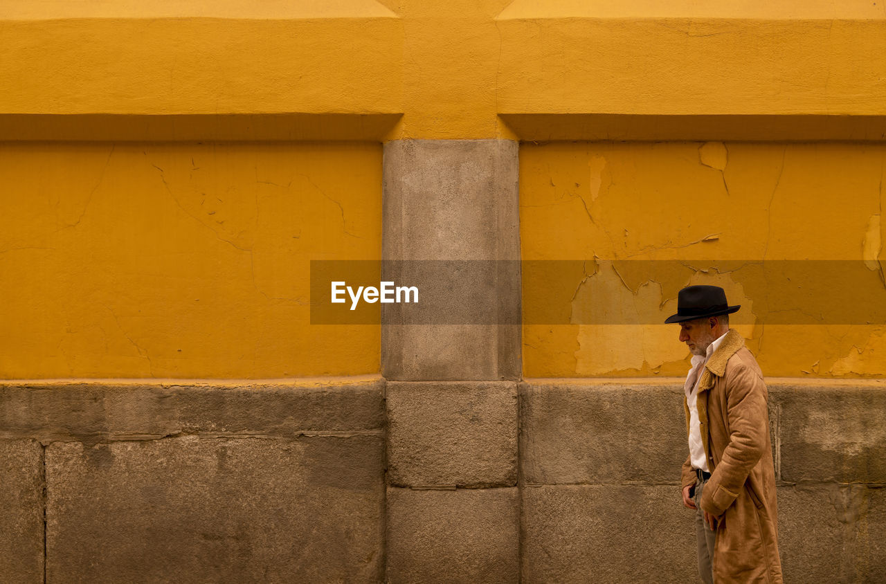 Adult man in hat and coat walking in front of yellow wall on street during sand storm day. madrid.