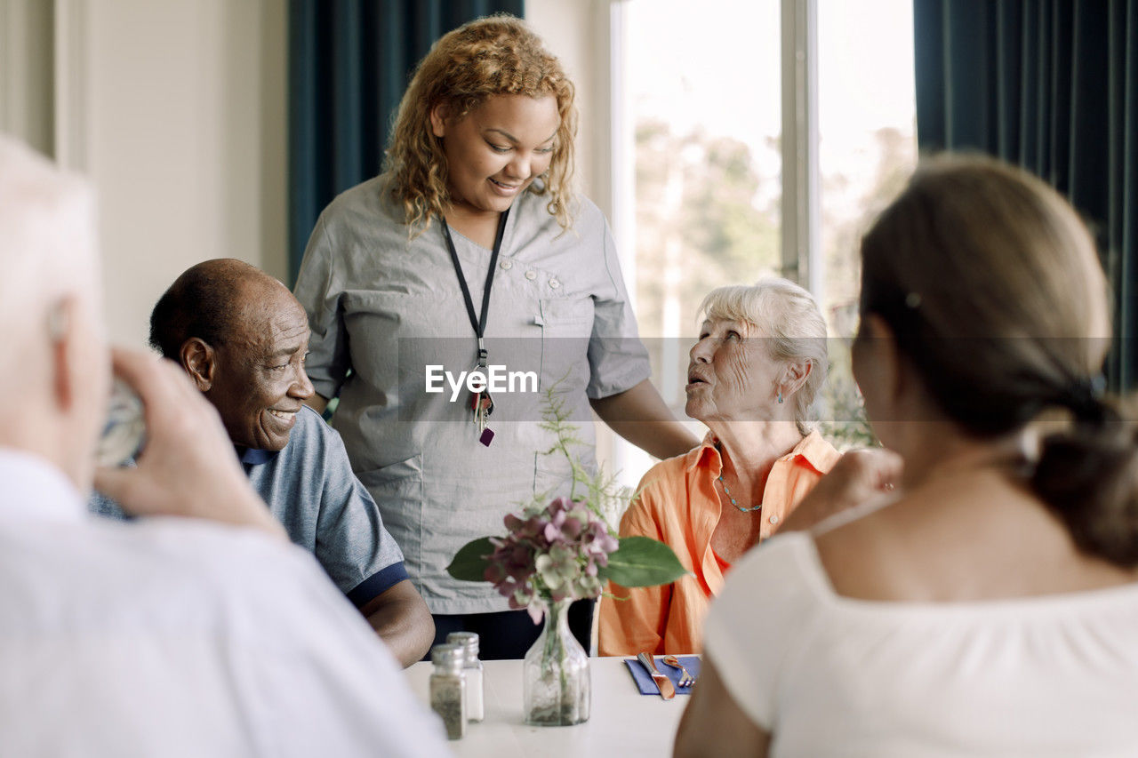 Smiling young female caregiver talking to senior women and men at dining table in nursing home