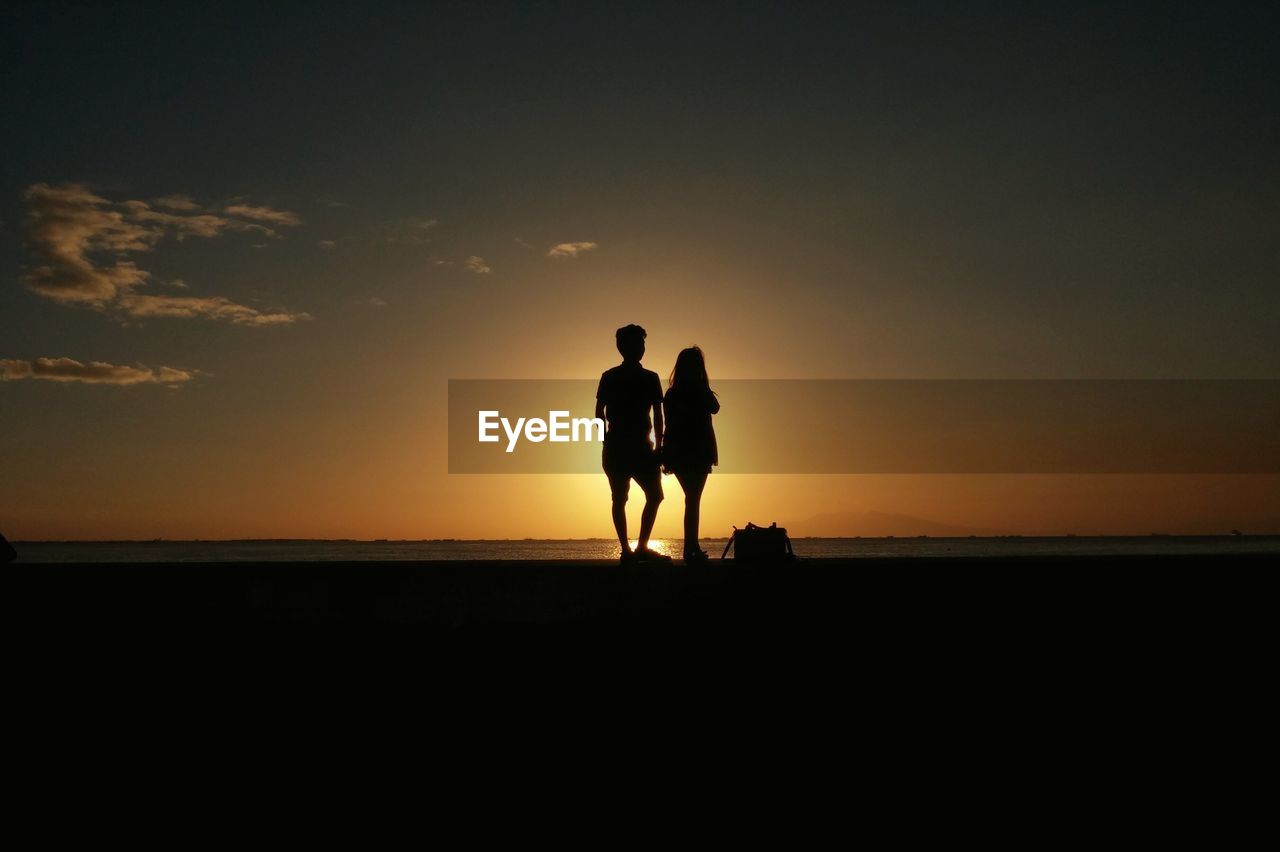 Silhouette friends standing at beach against sky during sunset