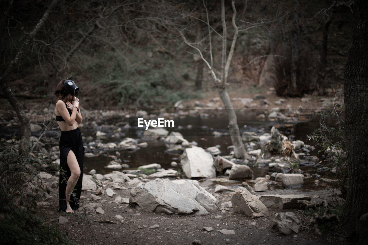 Side view of woman wearing gas mask while covering chest in forest