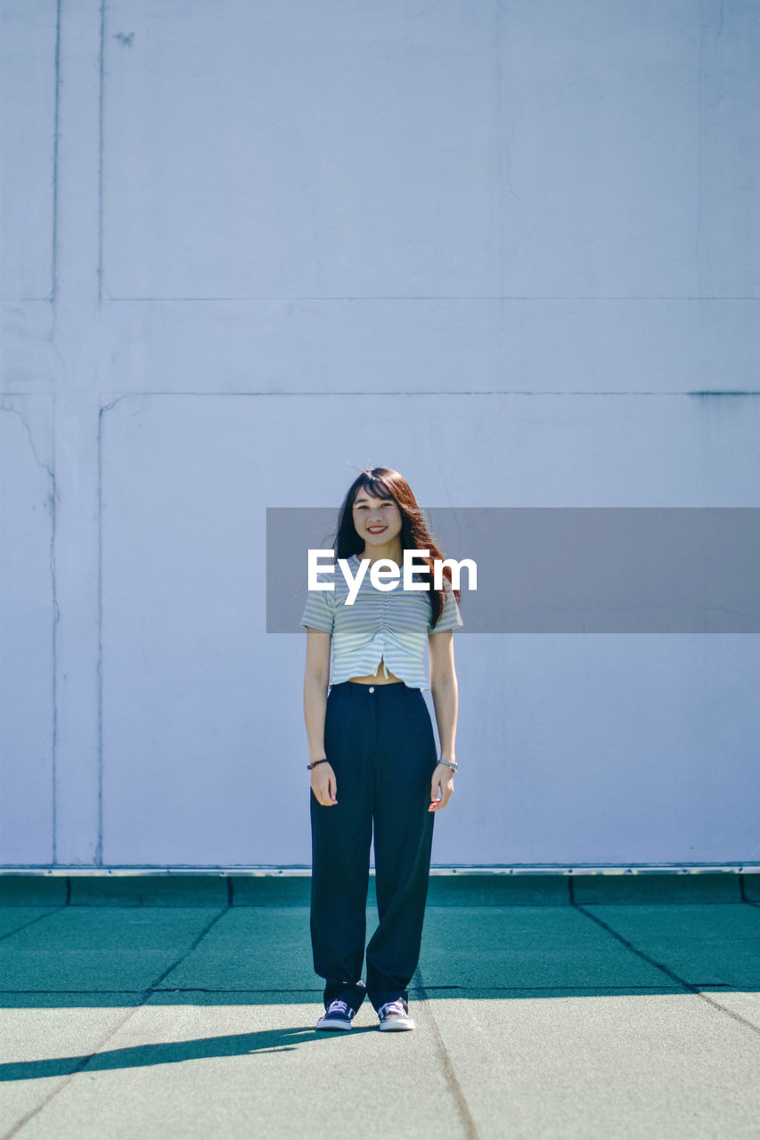 Full length of young woman smiling while standing against wall