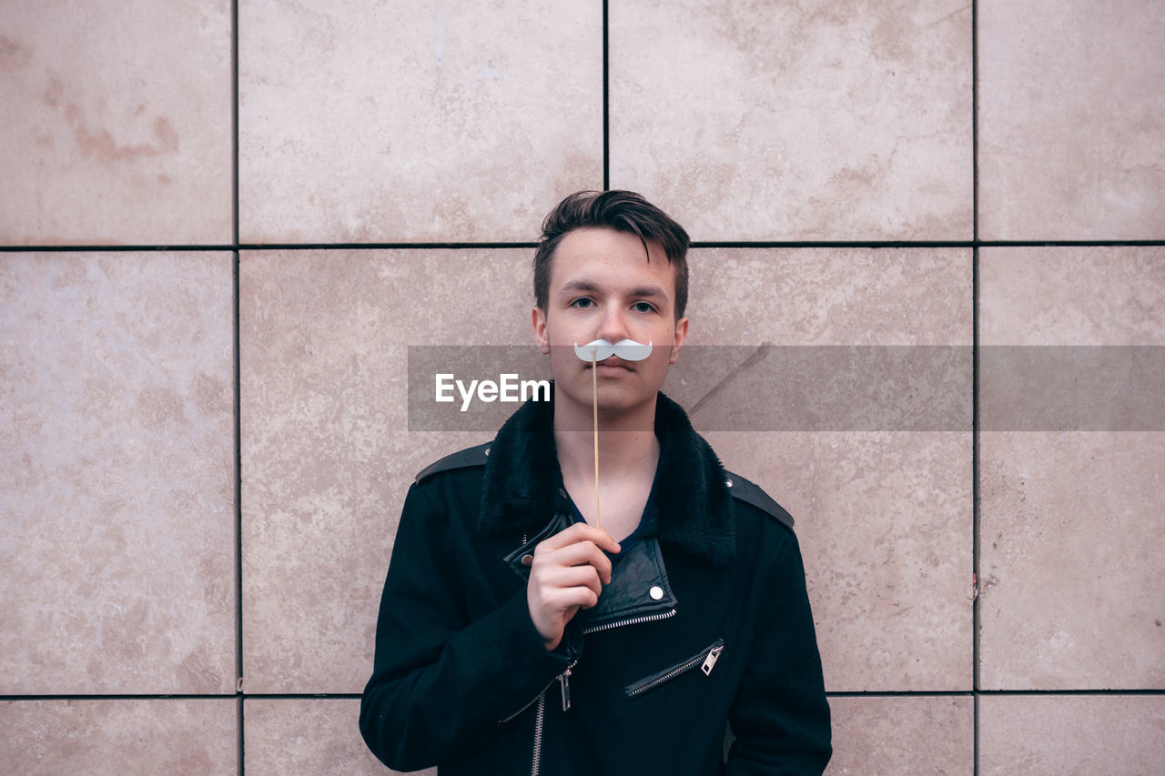 Portrait of young man holding mustache prop while standing against wall