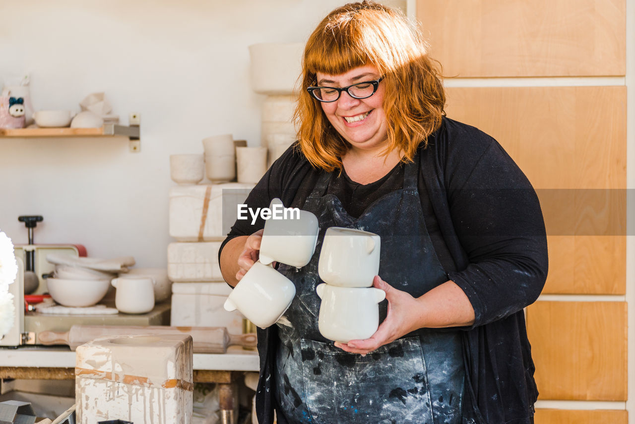 Smiling adult female artisan in dirty apron and black clothes standing in light studio and carrying handmade ceramic pots near clayware