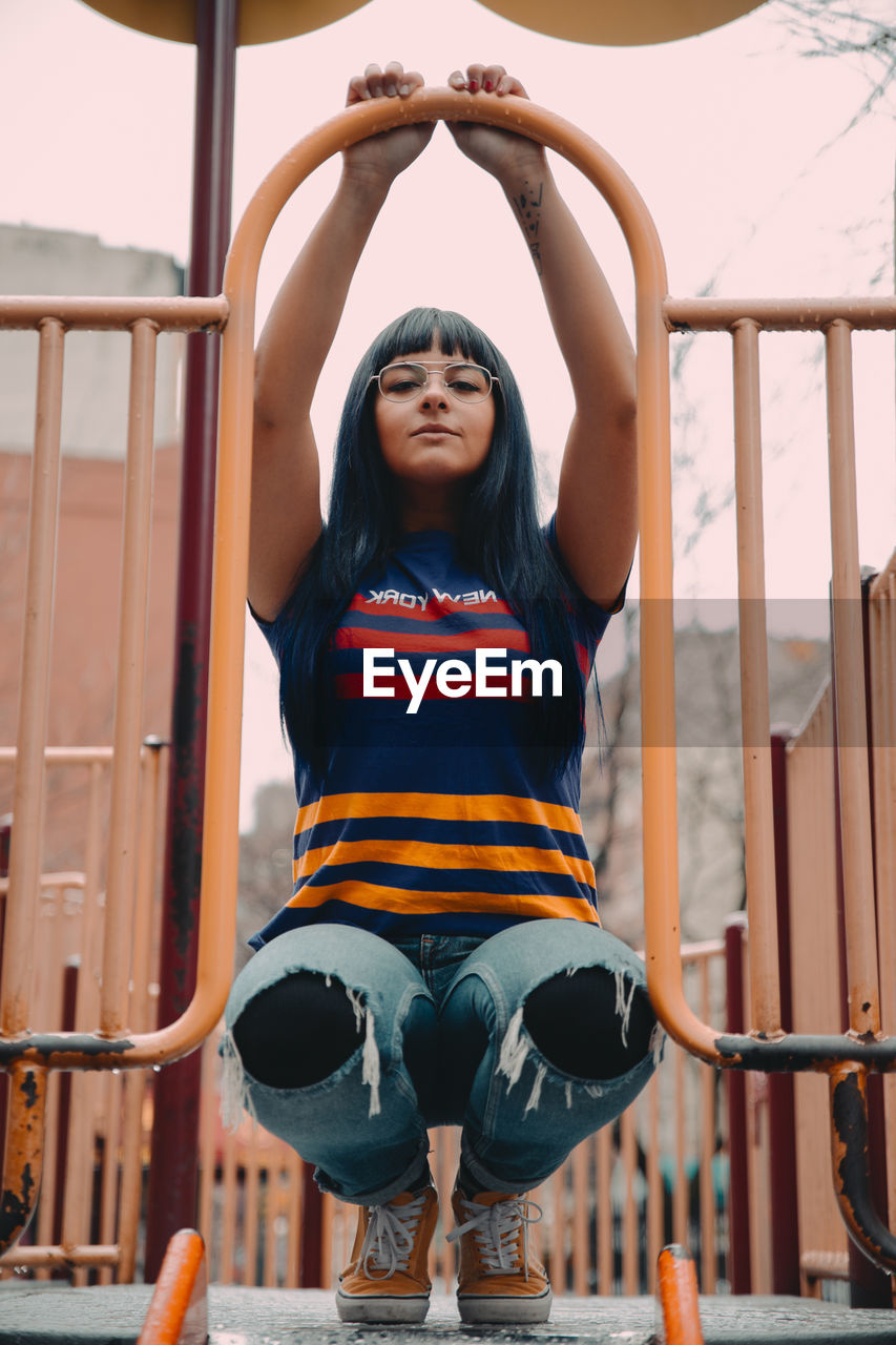 Full body of young ethnic female looking at camera while sitting on haunches on playhouse with metal fence while spending time on playground